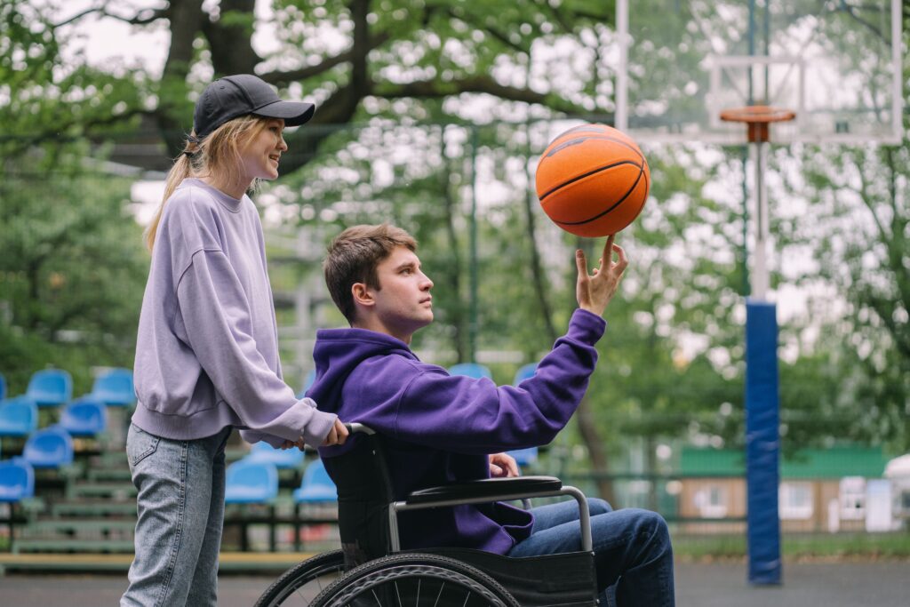 Twee tieners buiten, eentje in een rolstoel met een basketbal en de ander erachter